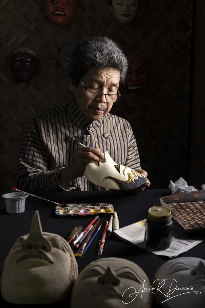 An elderly woman painting a decorative mask with gold accents at a table filled with art supplies and additional masks, with various masks hanging on a dark wall behind her.