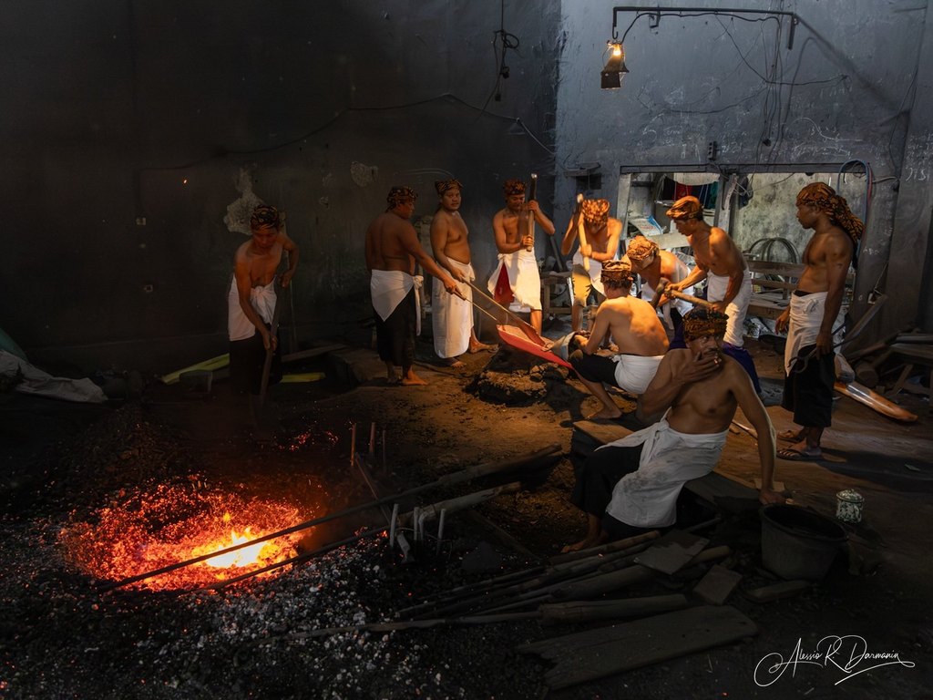 Traditional Indonesian artisans working with molten metal in a dark workshop, wearing headscarves and white cloths wrapped around their waists.