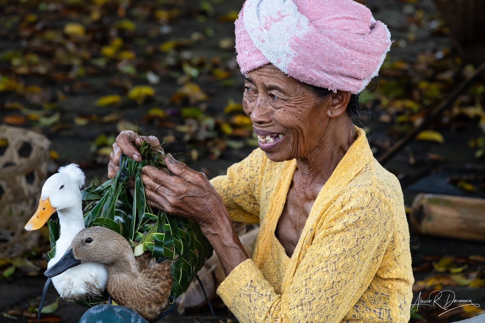 An elderly woman with a pink headwrap and yellow lace top crafting a bird decoration using real ducks' heads and green leaves outdoors.