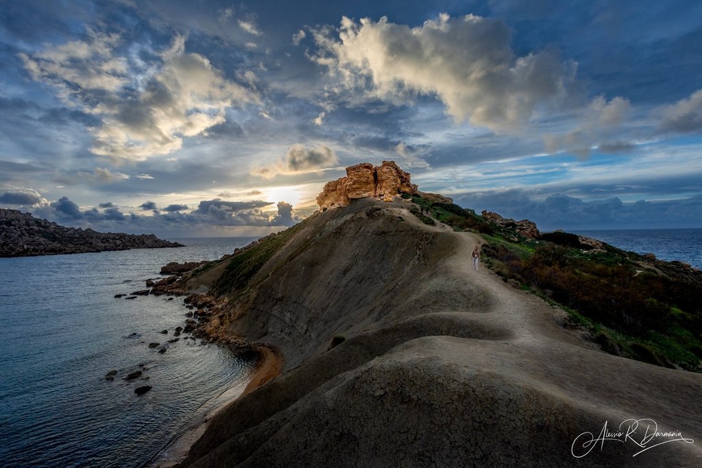 A coastal landscape features a dirt trail winding up a hill towards rocky formations at sunset, with the ocean on both sides and a sky filled with clouds.