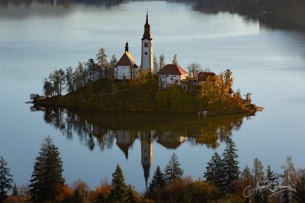 A small island with a church and houses surrounded by a calm lake, trees with autumn foliage, and a reflective shoreline.