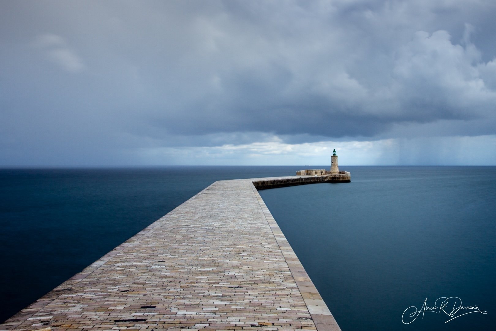A stone pier extending over calm ocean water toward a lighthouse on a cloudy sky day.