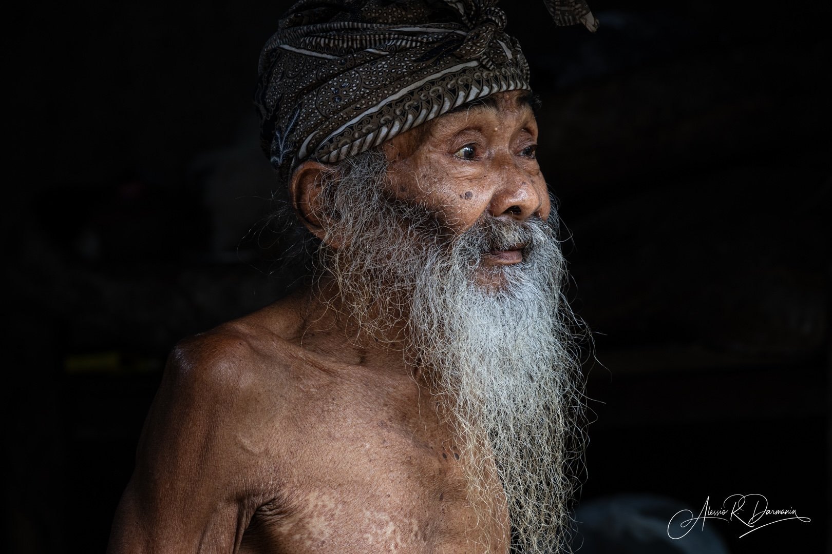 An elderly man with a long white beard, wearing a patterned headscarf, looking to the right with a calm expression, topless, against a dark background.