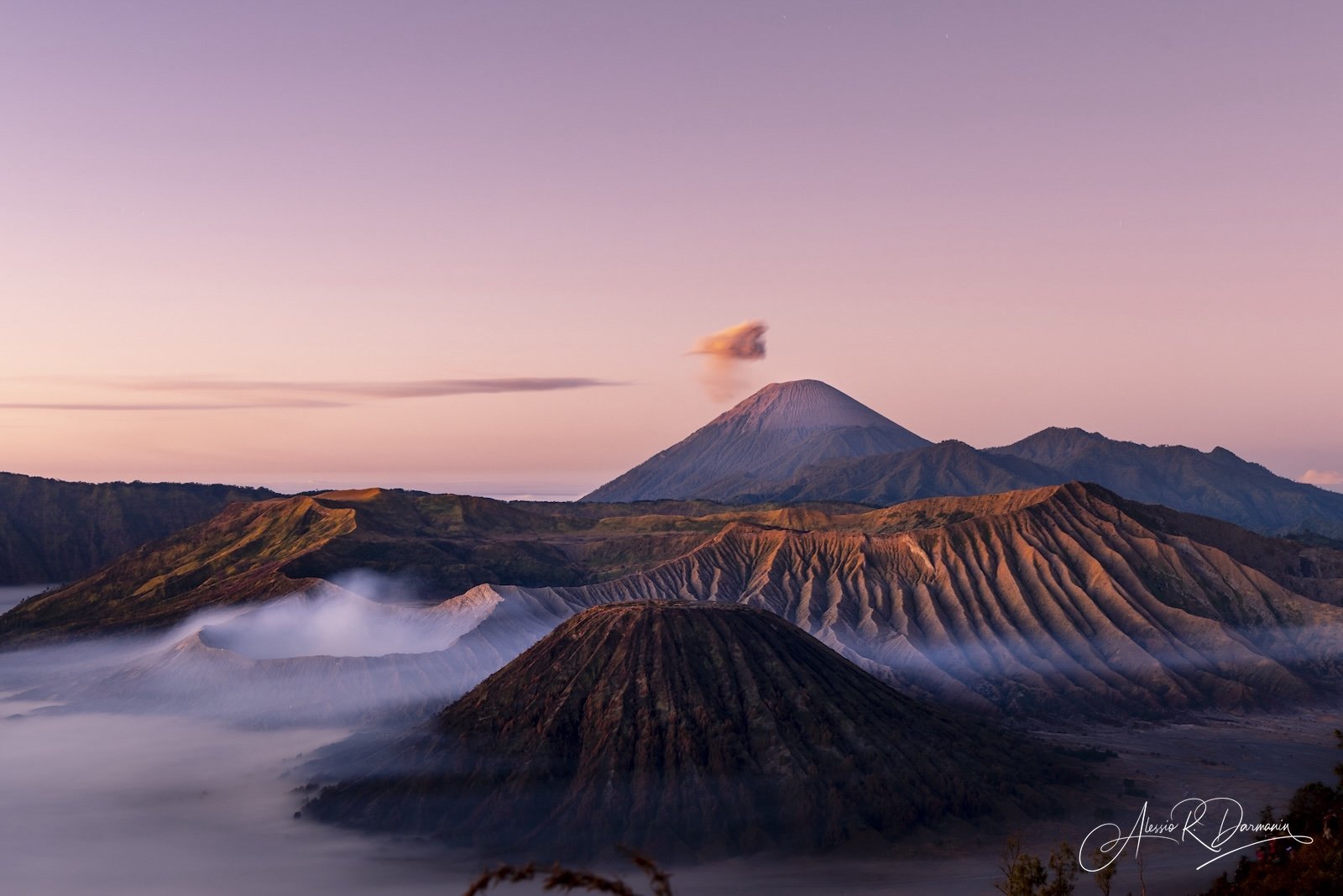 A volcanic landscape at sunrise with multiple volcanoes, one emitting a small plume of smoke, and colorful sky in the background.