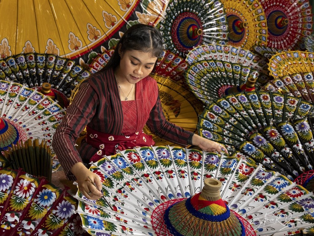 A woman in traditional attire painting colorful floral designs on a paper umbrella surrounded by multiple elaborately decorated umbrellas.
