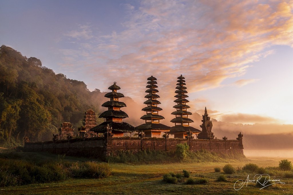 Traditional Balinese temple with layered thatched roofs, surrounded by lush greenery and misty hills at sunrise.