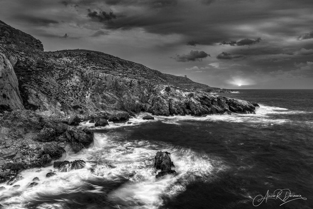 Black and white photograph of a rugged coastline with rocky cliffs and turbulent ocean waves crashing against the rocks, under a cloudy sky at sunset or sunrise.