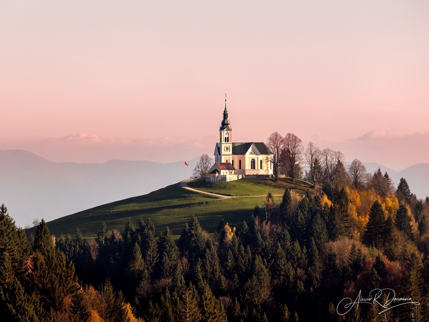 A white church with a tall steeple located on a hilltop surrounded by trees with autumn foliage, under a pink sky with distant mountains in the background.
