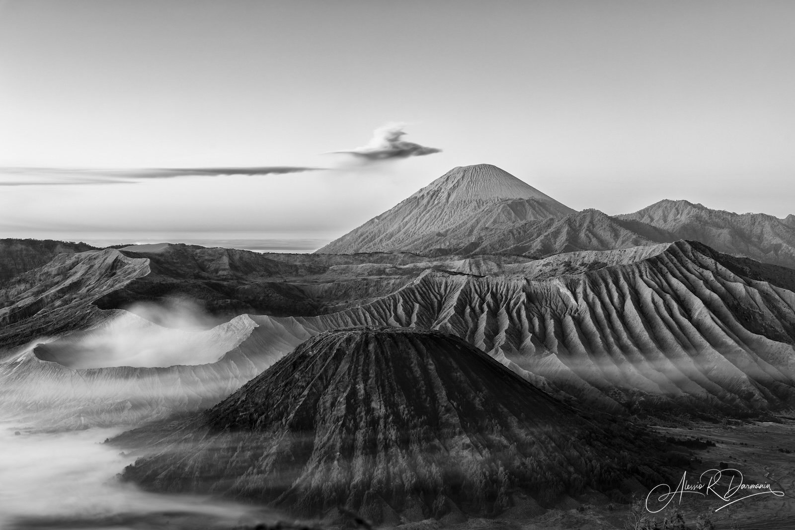 Black and white photograph of a volcanic landscape with mountains and smoke or steam rising from the crater, distant clouds, and rugged terrain.