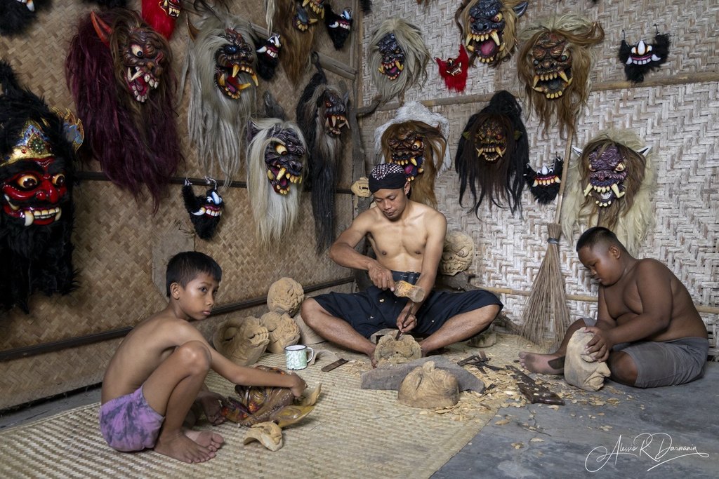 A man and two boys in a room surrounded by traditional masks and wooden sculptures, with the man carving a mask.