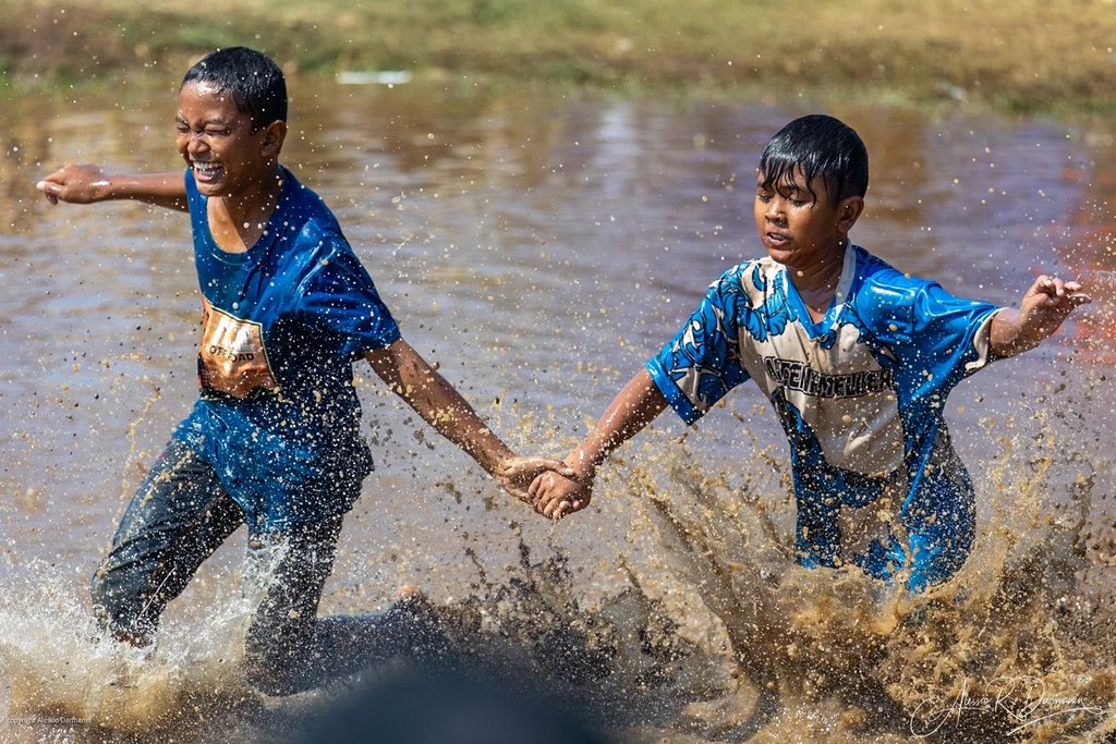 Two boys holding hands and playing in a muddy water puddle, splashing water around, with joyful expressions.