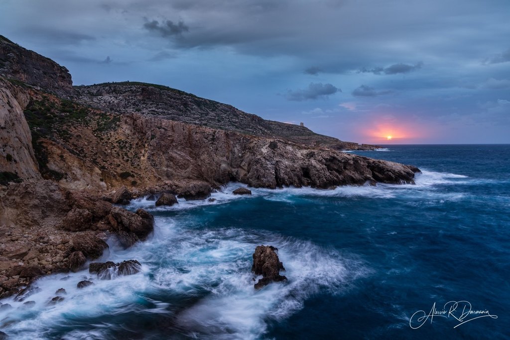 Scenic coastal view of rocky cliffs and the ocean during sunset or sunrise with waves crashing against rocks and a partly cloudy sky.