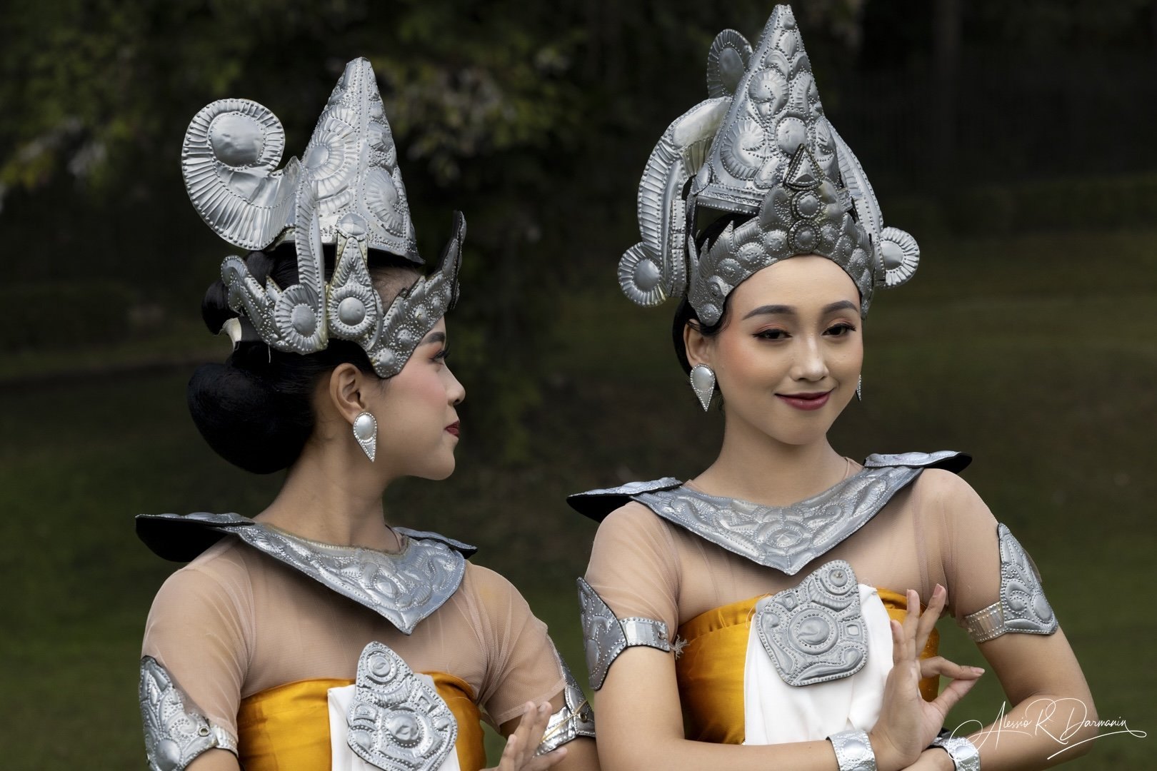 Two women in traditional costumes with intricate silver headpieces and jewelry, standing outdoors with a green background.