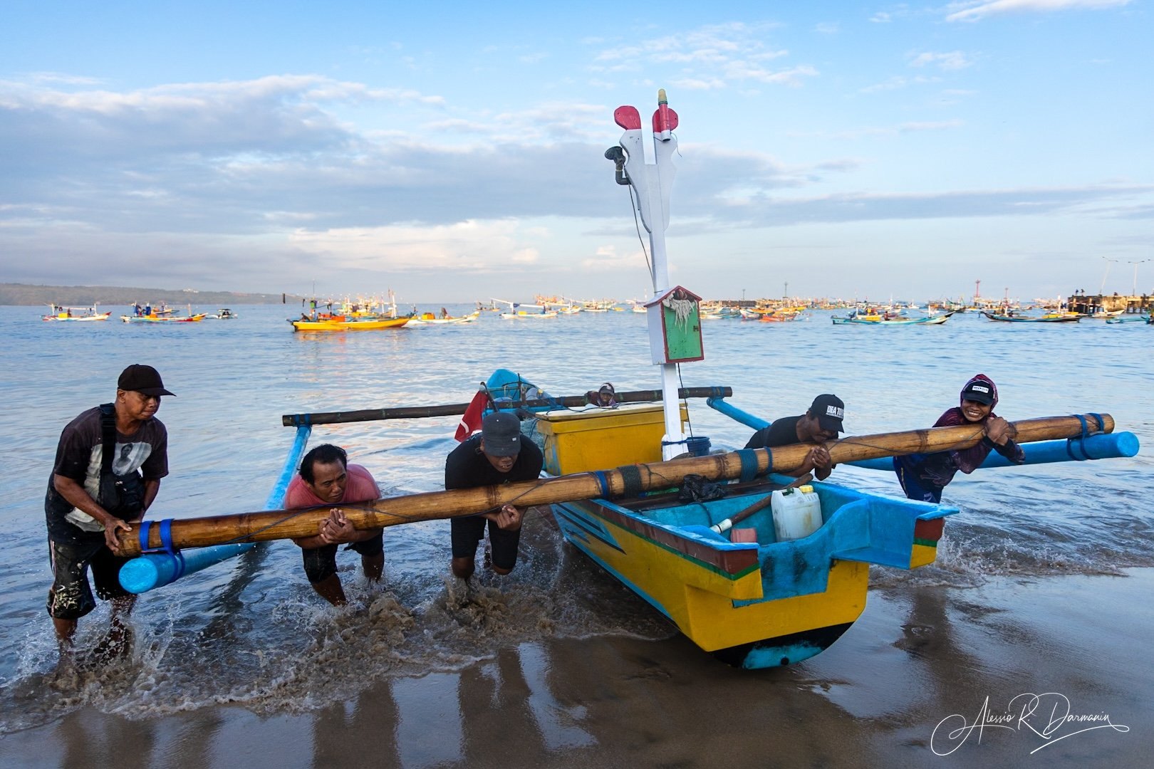 Four men are pushing a colorful small boat into the water at a beach, with numerous other boats on the water and a cloudy sky in the background.