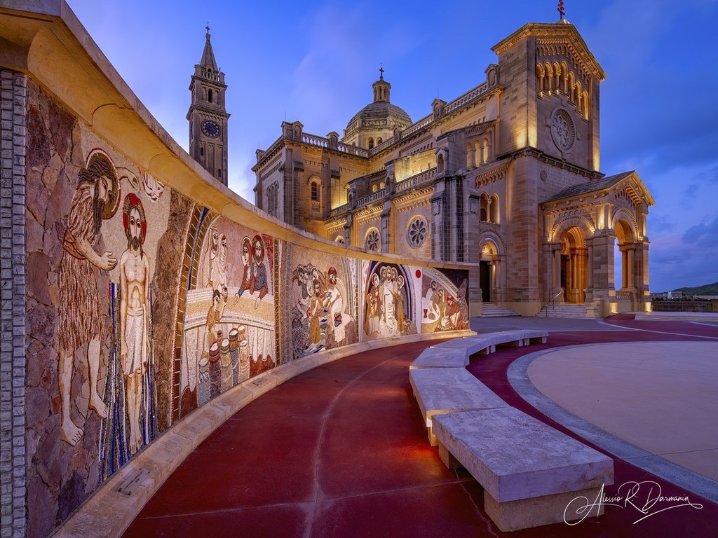 A historic church with illuminated architecture and a mosaic mural featuring biblical scenes along a red curved pathway at dusk.