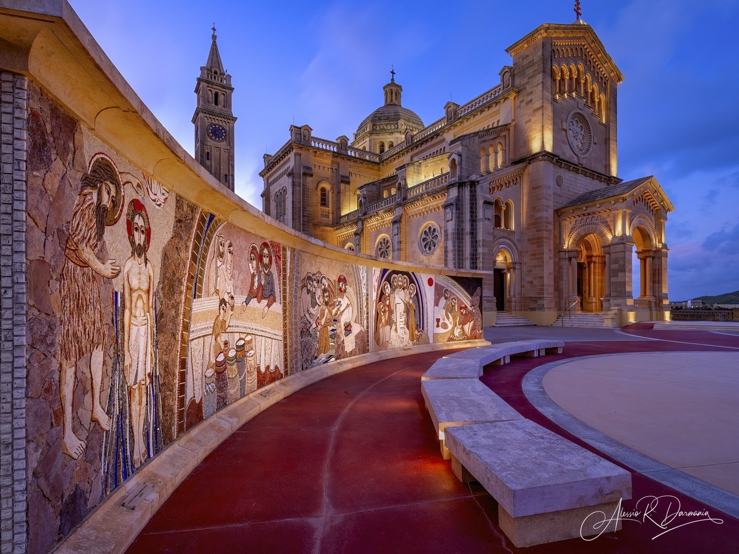 A historic church with illuminated architecture and a mosaic mural featuring biblical scenes along a red curved pathway at dusk.