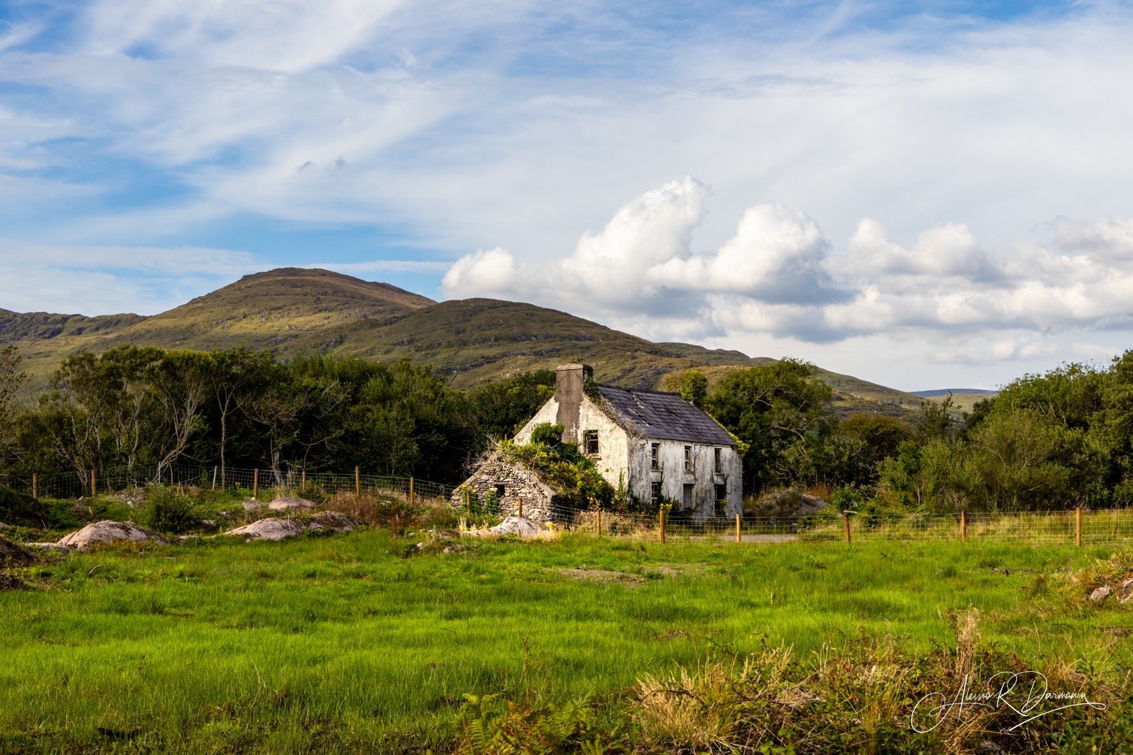 Old abandoned white house with broken windows surrounded by green grass and trees, with mountains and cloudy sky in the background.