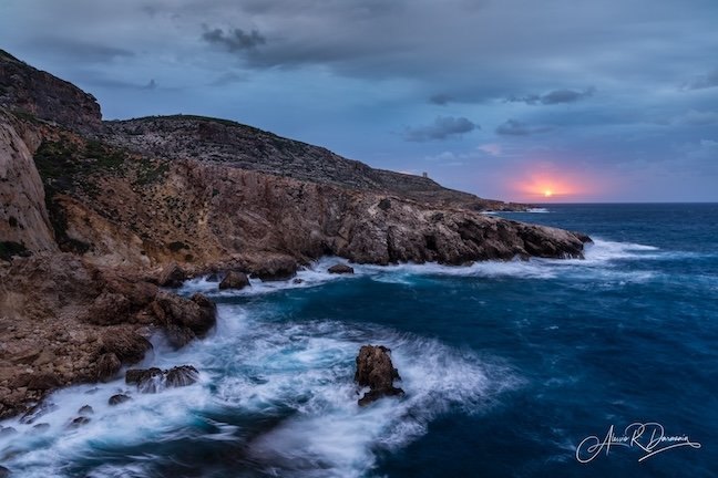 Scenic coastal view of rocky cliffs and the ocean during sunset or sunrise with waves crashing against rocks and a partly cloudy sky.