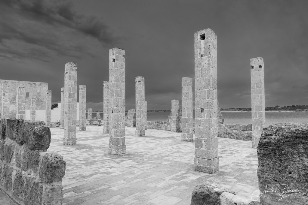 Ancient stone ruins with tall columns near a body of water, under a cloudy sky.